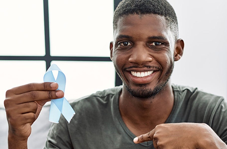 African American man wearing a moss green tee shirt and smiling as he holds up a blue ribbon which represents surviving prostate cancer