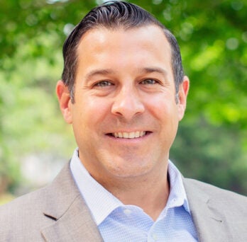 Man with short dark hair wearing a light blue shirt and gray suit coat, posing amidst trees on a sunny day and smiling into the camera 