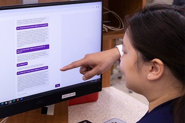 a young woman wearing with long dark hair and wearing dark blue scrubs sitting at a computer and interacting with the chat bot