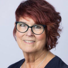 woman with dark red hair and glasses and wearing a black blouse, smiling as she poses in front of a gray backdrop