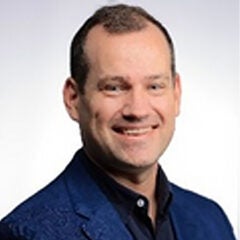 Man with short dark hair wearing a black shirt and dark blue suit coat, posing against a gray backdrop and smiling into the camera