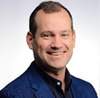 Man with short dark hair wearing a black shirt and dark blue suit coat, posing against a gray backdrop and smiling into the camera 