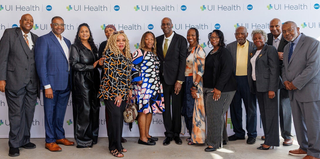 Thirteen African American men and woman in formal attire, smiling as they pose together in front of a backdrop of UI Health logos