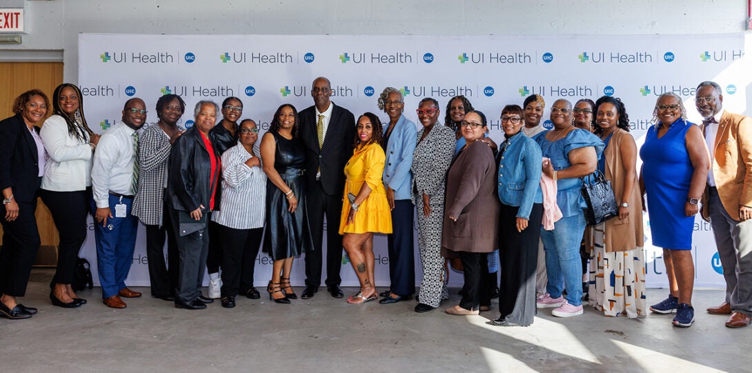 Twenty-one African American men and woman in formal attire, smiling as they pose together in front of a backdrop of UI Health logos