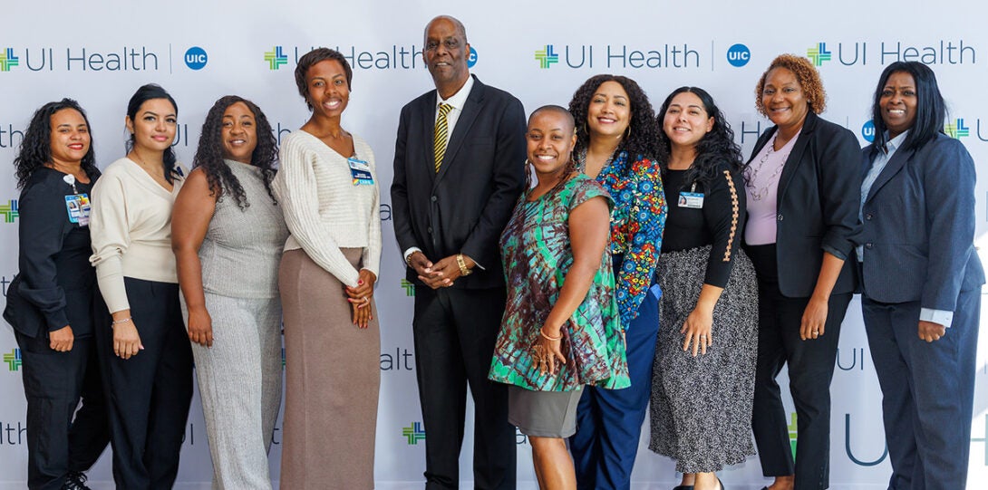 A diverse group of ten people of color in formal attire, smiling as they pose together in front of a backdrop of UI Health logos