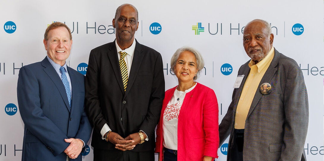 : A white man, a Black man, a Latinx woman and another Black man, smiling as they pose together in front of a backdrop of UI Health logos. The mean are wearing suit coats and the woman is wearing a bright red jacket.