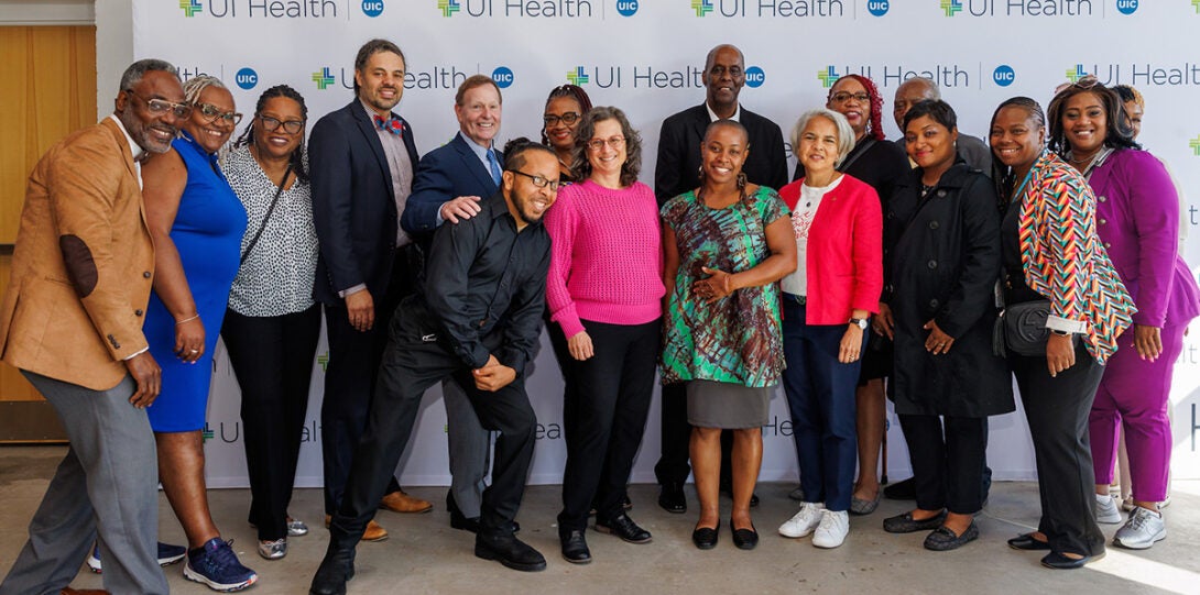 : A diverse group of seventeen people, smiling as they pose together in front of a backdrop of UI Health logos