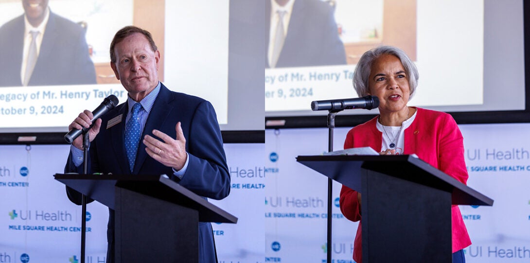 At left a white man wearing a dark blue suit, striped shirt and blue necktie, at right a Latinx woman wearing a white top and bright red jacket, both standing at a podium and speaking into a microphone