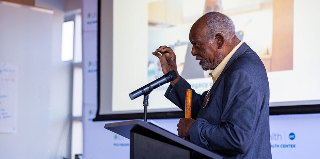 An older African American man wearing a blue suit and yellow shirt, standing at a podium and speaking into a microphone, gesturing with his hand as he speaks