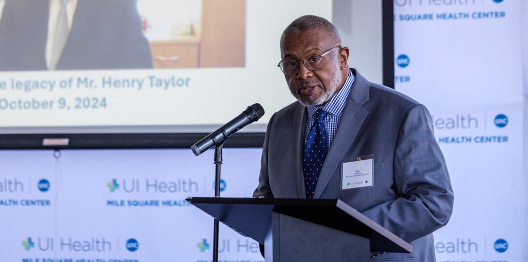 An older African American man wearing a gray suit, checked shirt and blue necktie, standing at a podium and speaking into a microphone