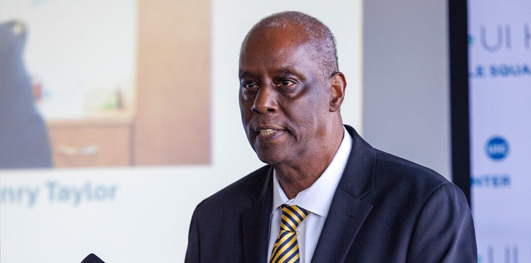 : An older African American man wearing a dark blue suit, white shirt and striped necktie, looking out at an audience as he is speaking