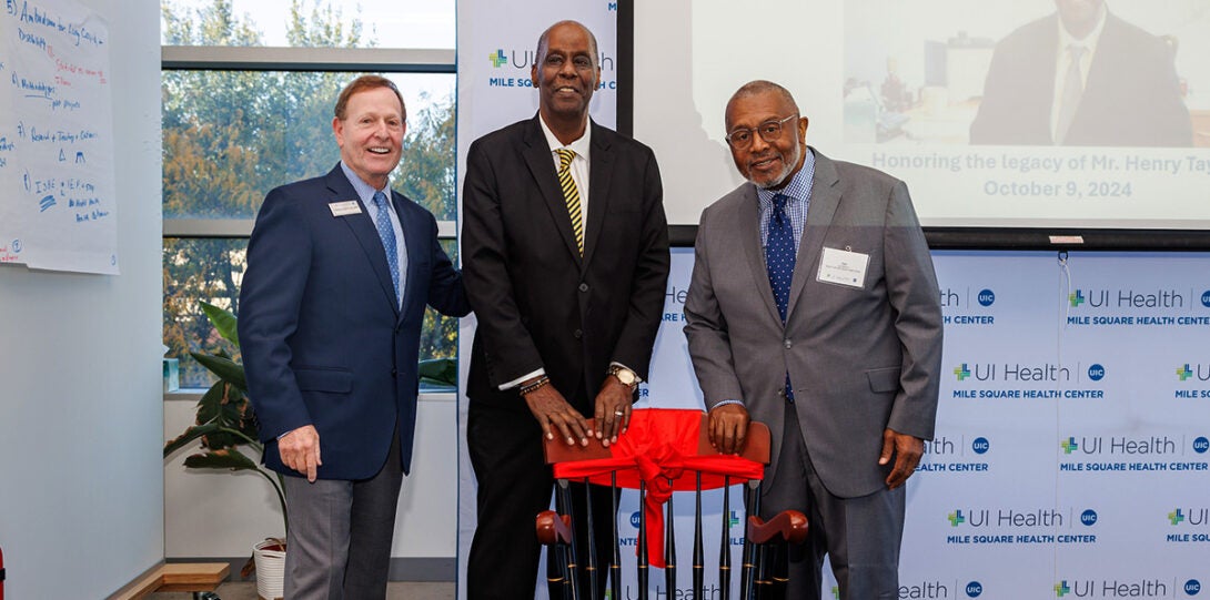 : A white man and two African American men, smiling and posing together behind a wooden captain’s chair that is adorned with a large red ribbon