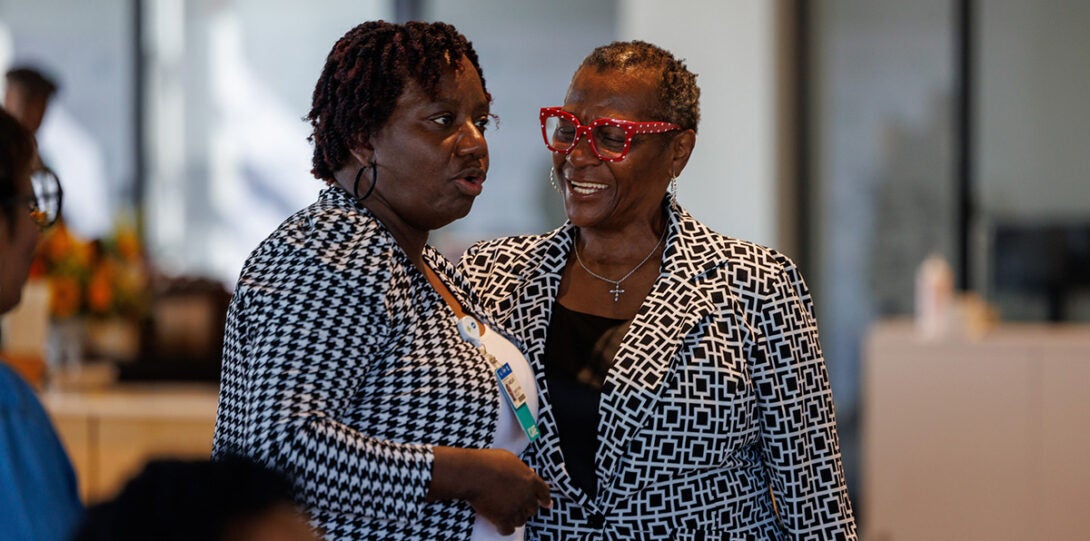 Two African American, both wearing black and white patterned jackets and one wearing red eye glasses, smiling as they converse