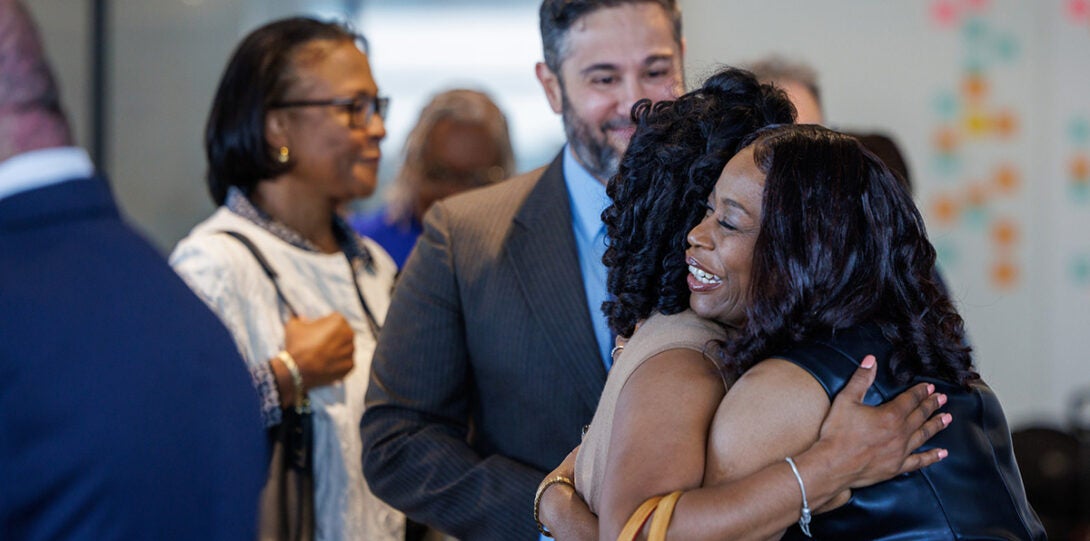 Two African American women hugging with a diverse group of people in the background