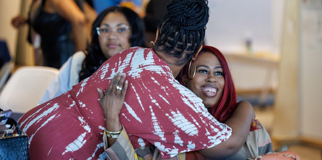 An African American women in a red and white dress, hugging another African American woman who has red-tinted hair