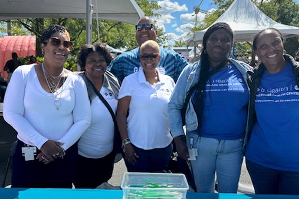 Six people standing behind a table covered with information outside on a sunny day.