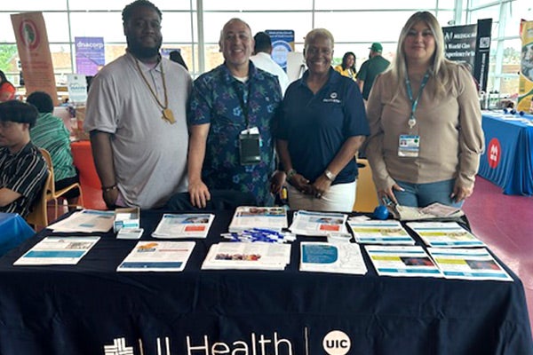 Four people standing behind a table in an event space. The table has a dark blue UI Health table cloth and is covered with flyers and information.