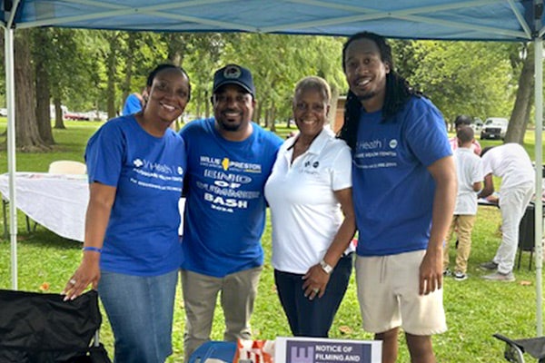 Four people, three wearing UI Health t shirts, standing behind a table under a low tent in a park