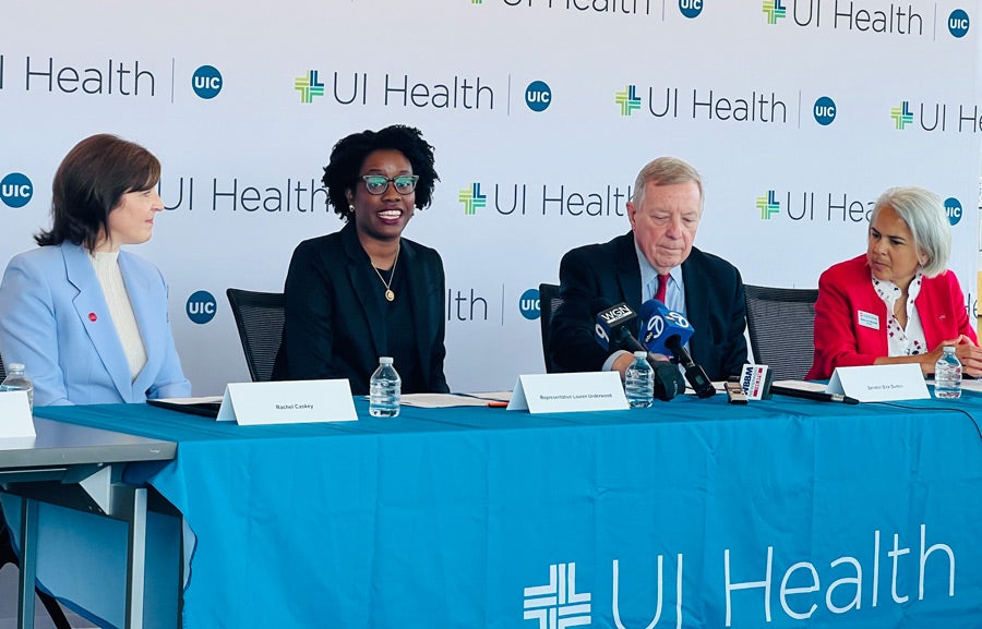 The people named in the caption, all sitting behind a table with a blue UI Health table cloth. Representative Lauren Underwood is speaking. Behind them is a background of repeating UI Health logos.