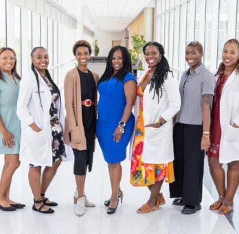 Nine African American women standing in a well-lit clinical hallway, some wearing formal attire and some wearing white lab coats. 