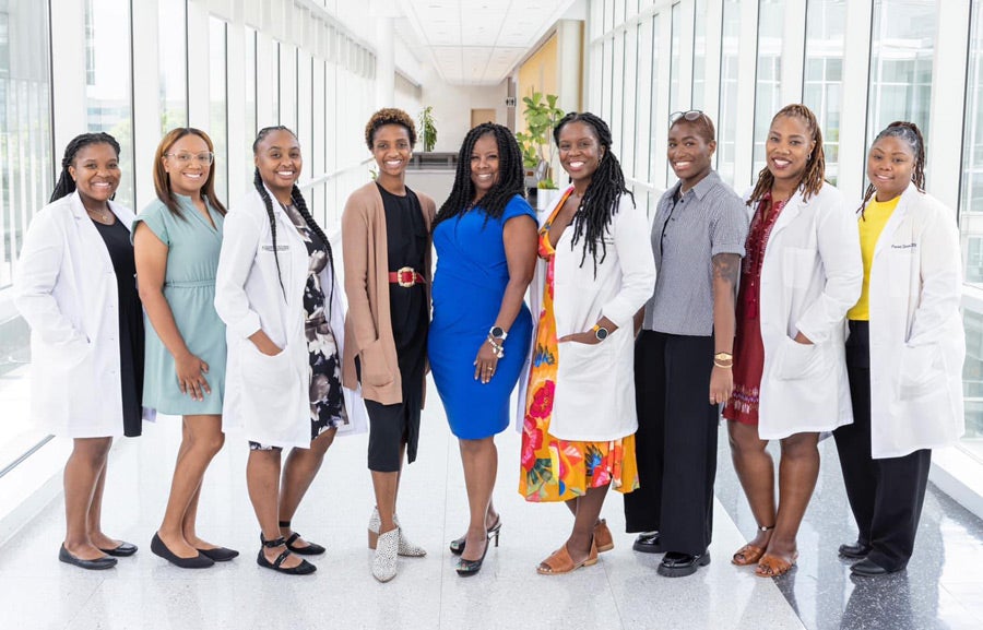 Nine African American women standing in a well-lit clinical hallway, some wearing formal attire and some wearing white lab coats.