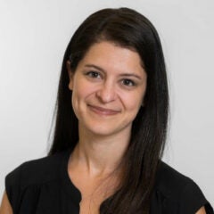 woman with long dark hair and wearing a black dress, posed in front of a gray backdrop and and smiling into the camera