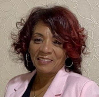 Black woman with shoulder-length red-tinted hair, wearing a pink jacket and smiling into the camera 