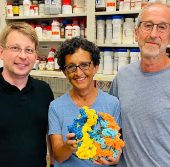 A young man with blond hair, a woman with dark hair and glasses, and a man with glasses beard and mustache, posing together in a laboratory setting 