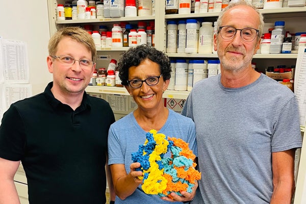 A young man with blond hair, a woman with dark hair and glasses, and a man with glasses beard and mustache, posing together in a laboratory setting