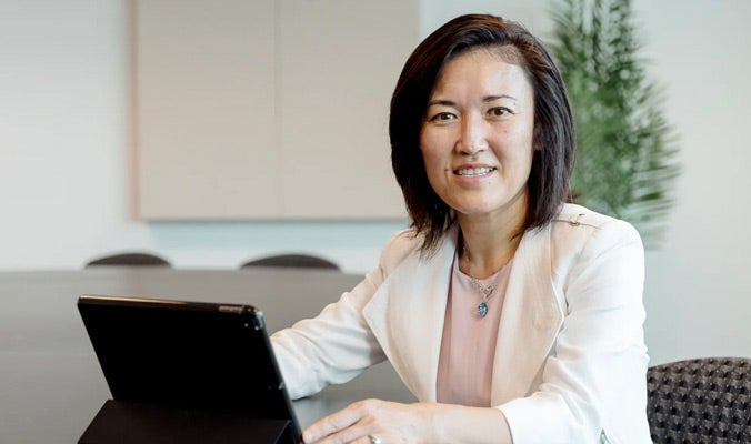 An Asian woman with shoulder length dark hair, wearing a white jacket and pink blouse, sitting at a desk with a laptop computer Dr. Jun Ma, Beth Fowler Vitoux and George Vitoux Distinguished Professor in Geriatrics at the College of Medicine.