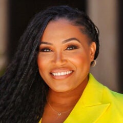 Black woman with long braided hair, wearing a yellow suit jacket and smiling into the camera