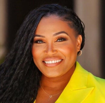 Black woman with long braided hair, wearing a yellow suit jacket and smiling into the camera 