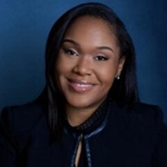 Black woman with long hair, wearing a dark blue suit jacket and smiling into the camera