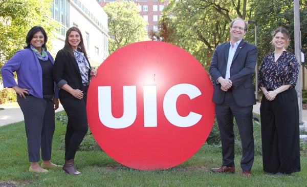 Four UIC faculty members smiling as they pose outside on a sunny day, next to a large red UIC sign