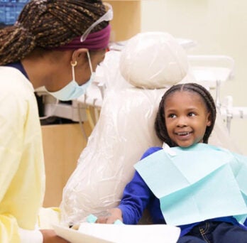 Young African American girl sitting in a dental chair and smiling as dentist in yellow scrubs speaks with her 