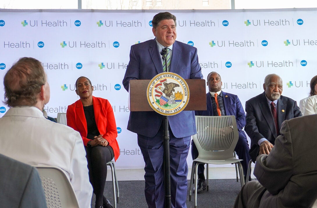 Governor Pritzker on a stage behind a podium, with other legislators seated behind him